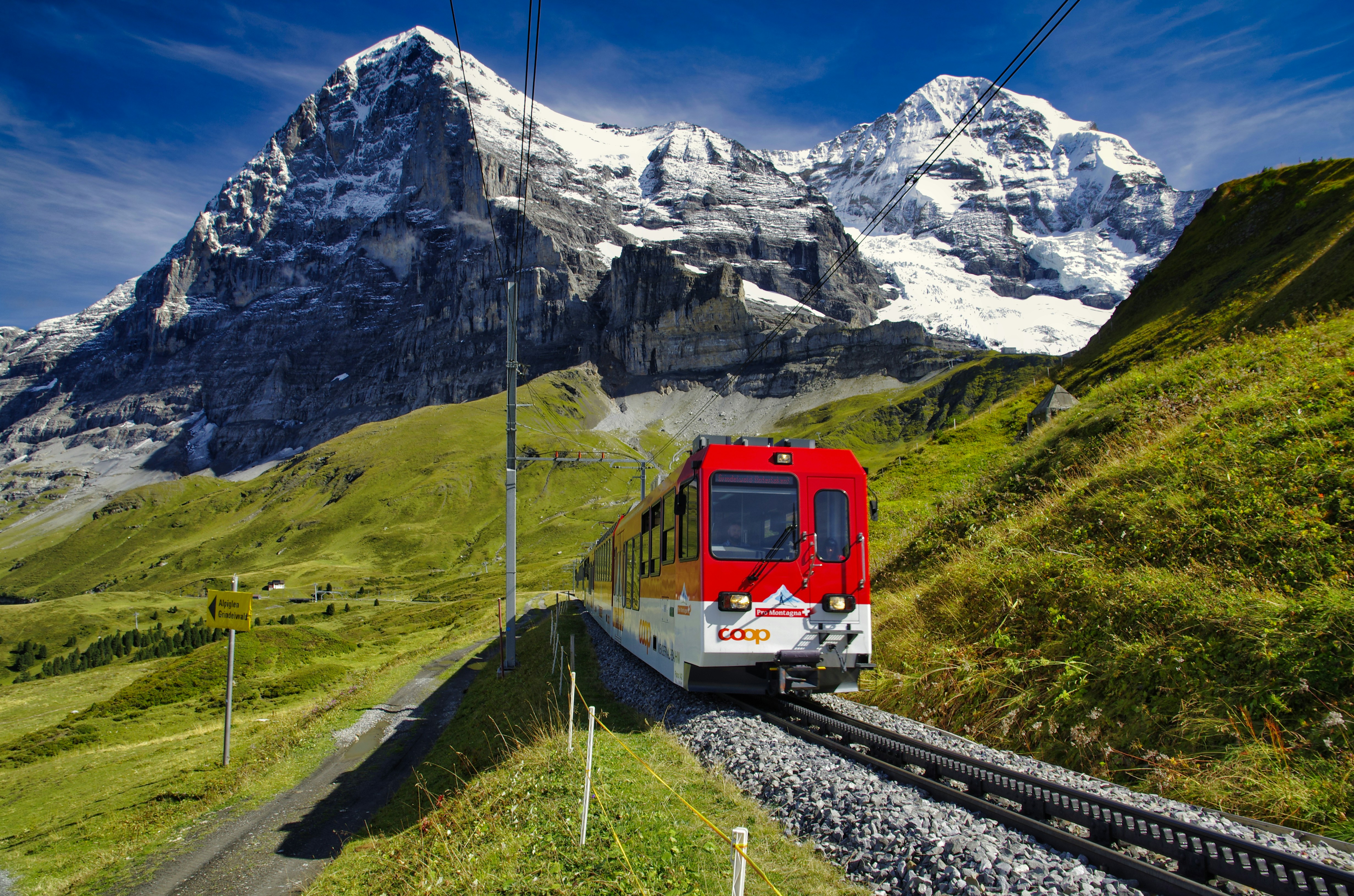 An illustrative photo of a red and white train on a rail road near a snow covered mountain.