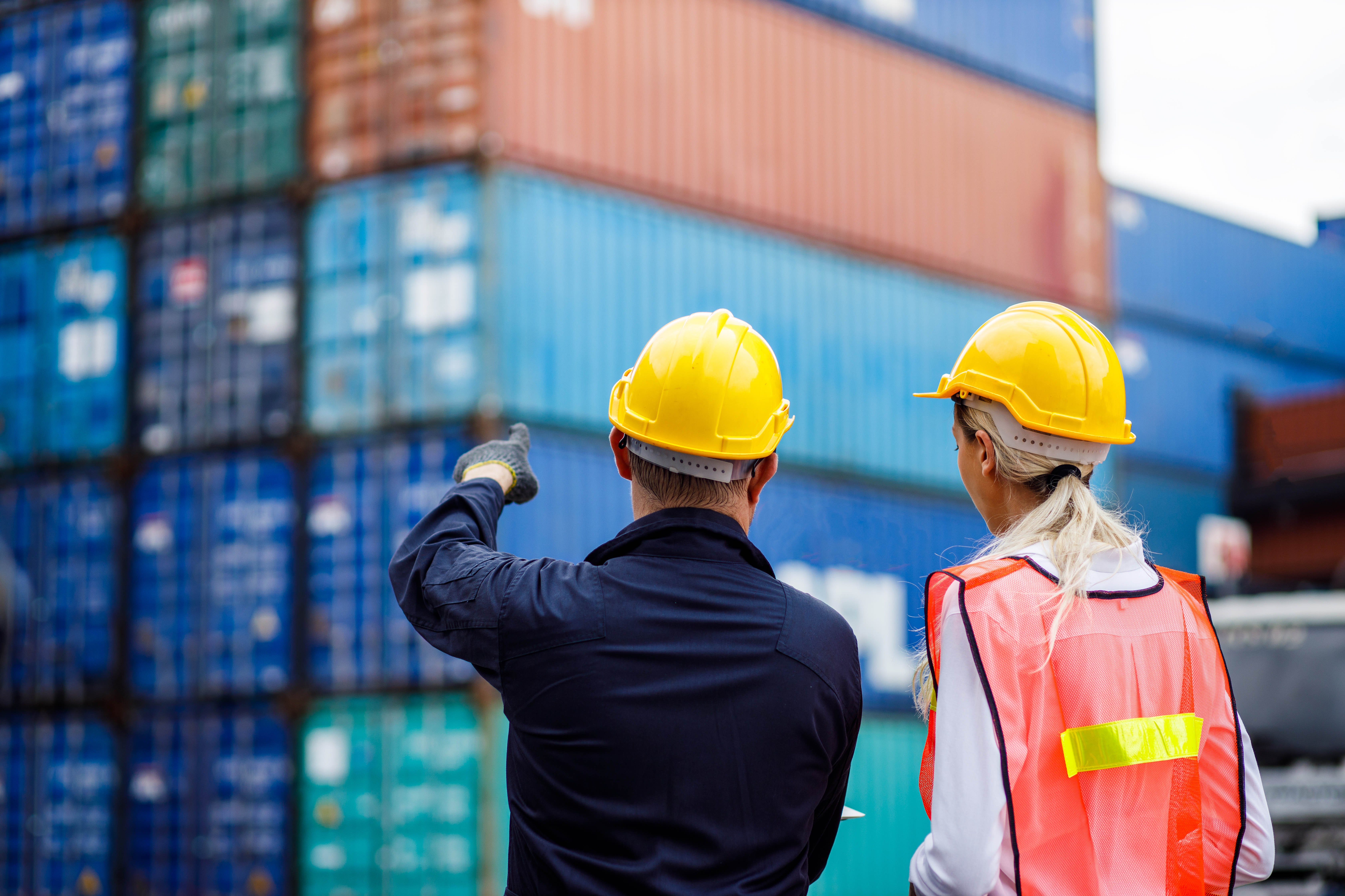 A photo presenting two workers in safety helmets inspecting stacked shipping containers at a cargo terminal.