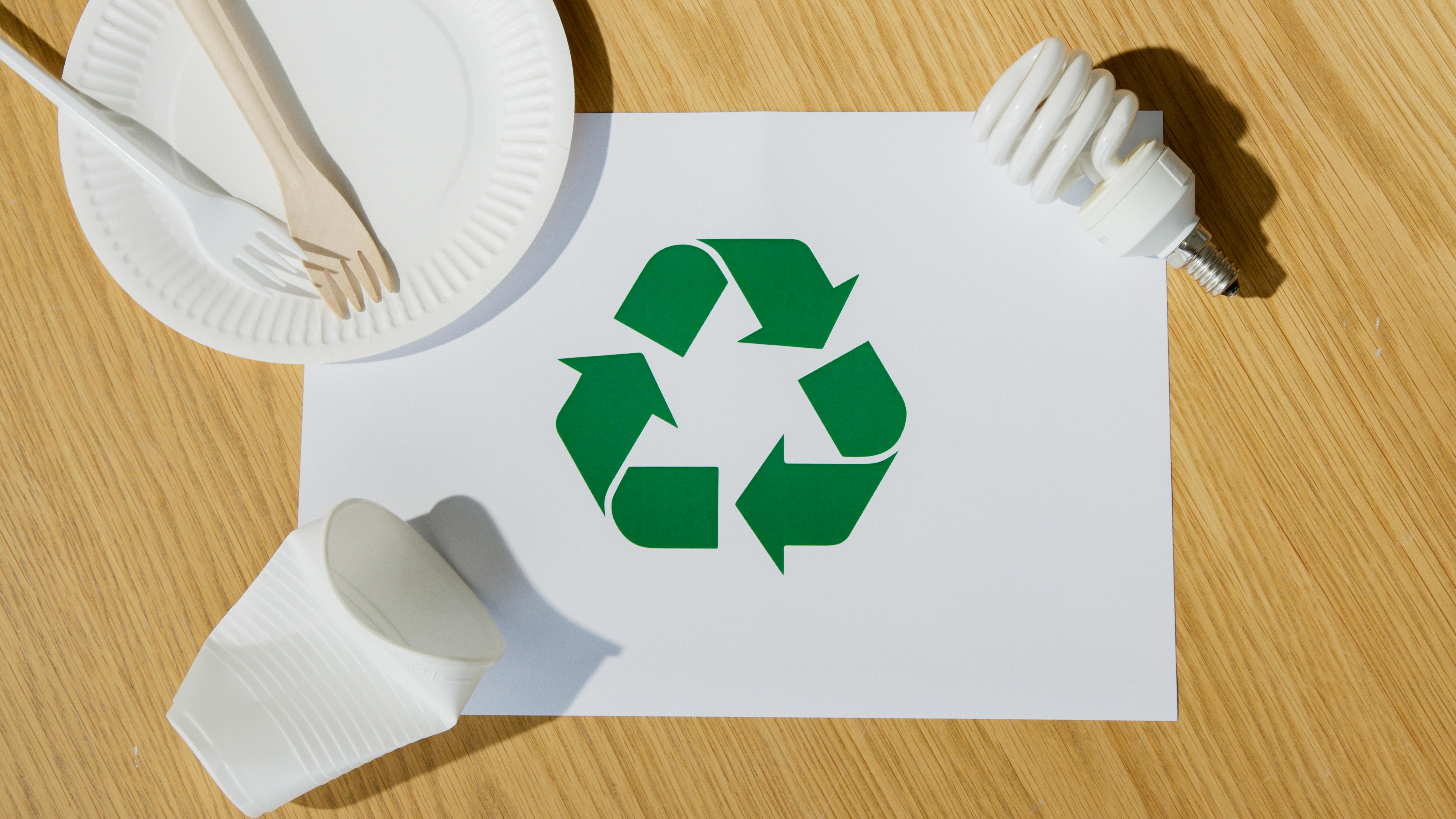 Recycling symbol on paper surrounded by disposable tableware and an energy-saving light bulb on a wooden surface.