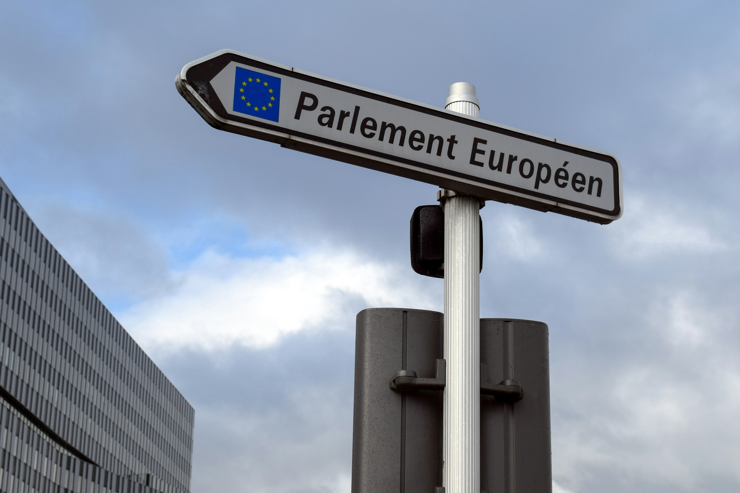 Street sign pointing to the European Parliament, with the EU flag visible against a cloudy sky.