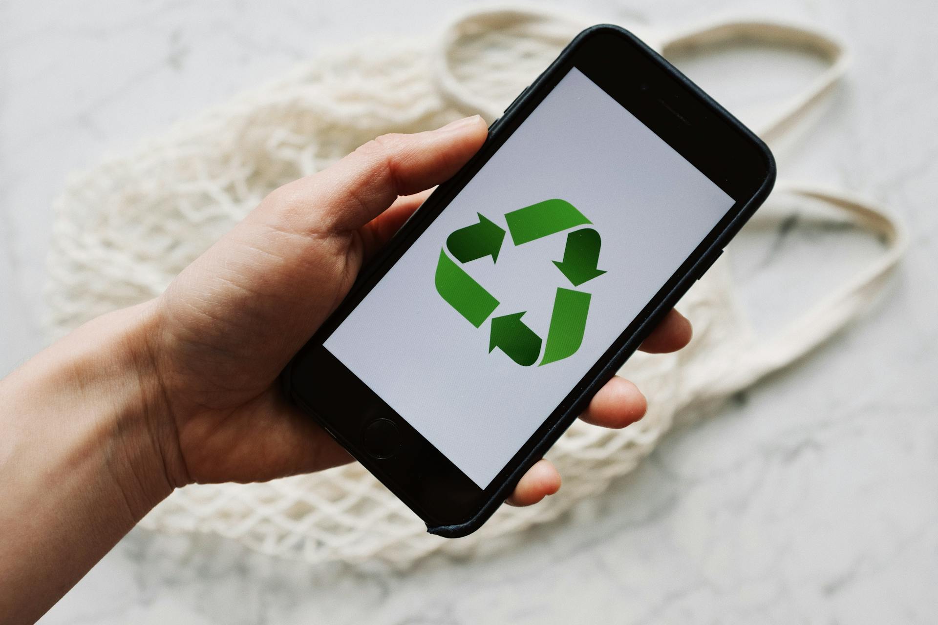 A hand holding a smartphone displaying a green recycling symbol, with a reusable mesh bag in the background.