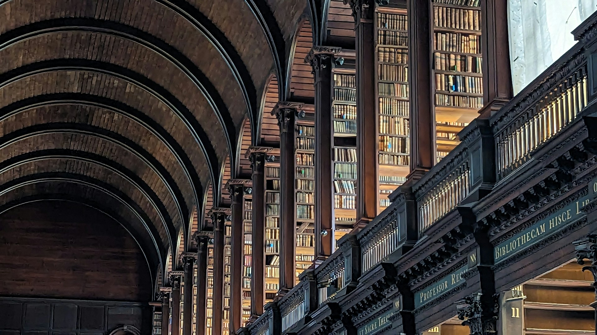 The image shows a grand library interior, with tall wooden bookshelves stretching into the distance and high arched ceilings. Rows of books are neatly arranged along both sides of a long hall. The architecture and layout suggest a historic European library, possibly in an academic setting.