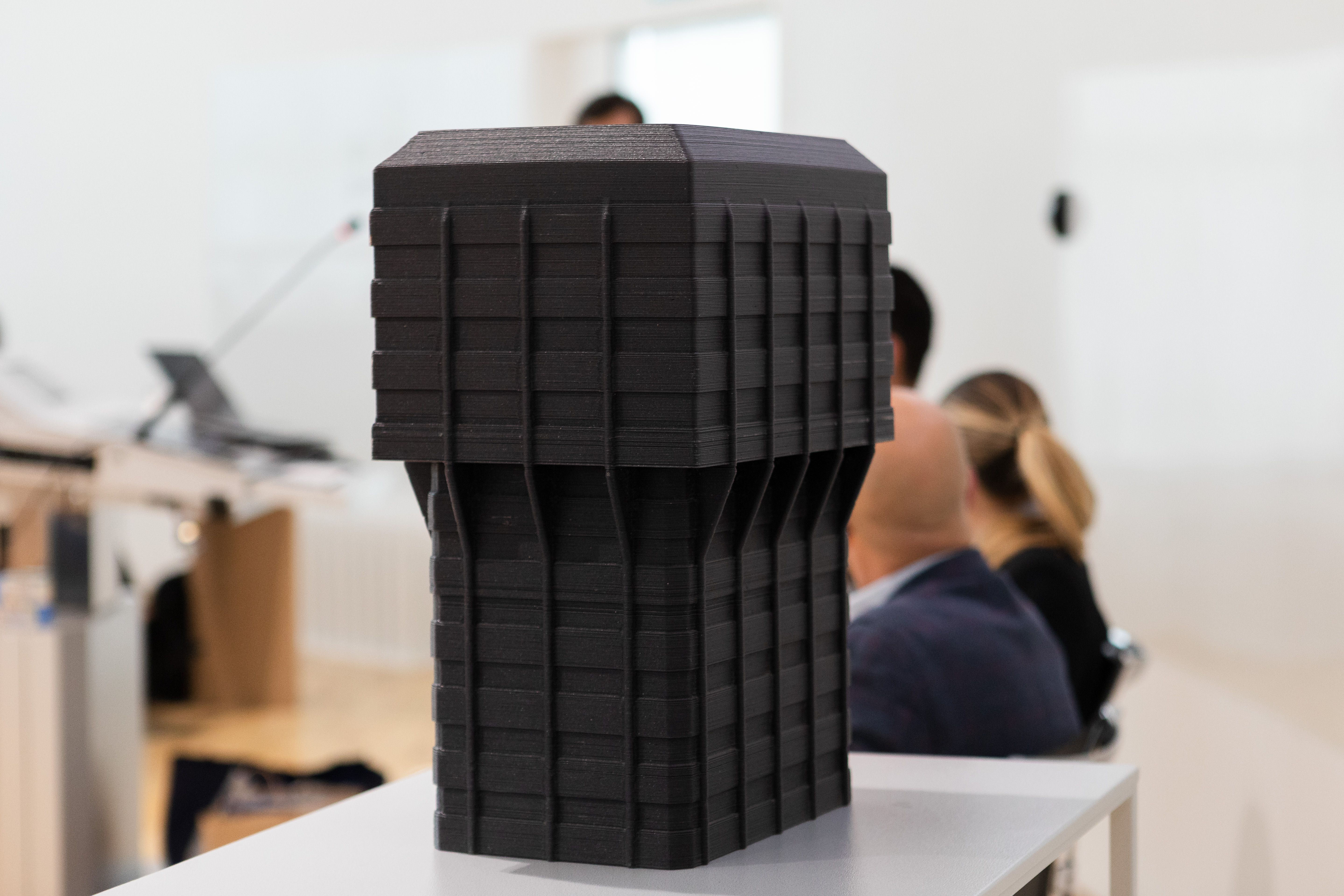 Layered 3D-printed object displayed on a table, with people blurred in the background.