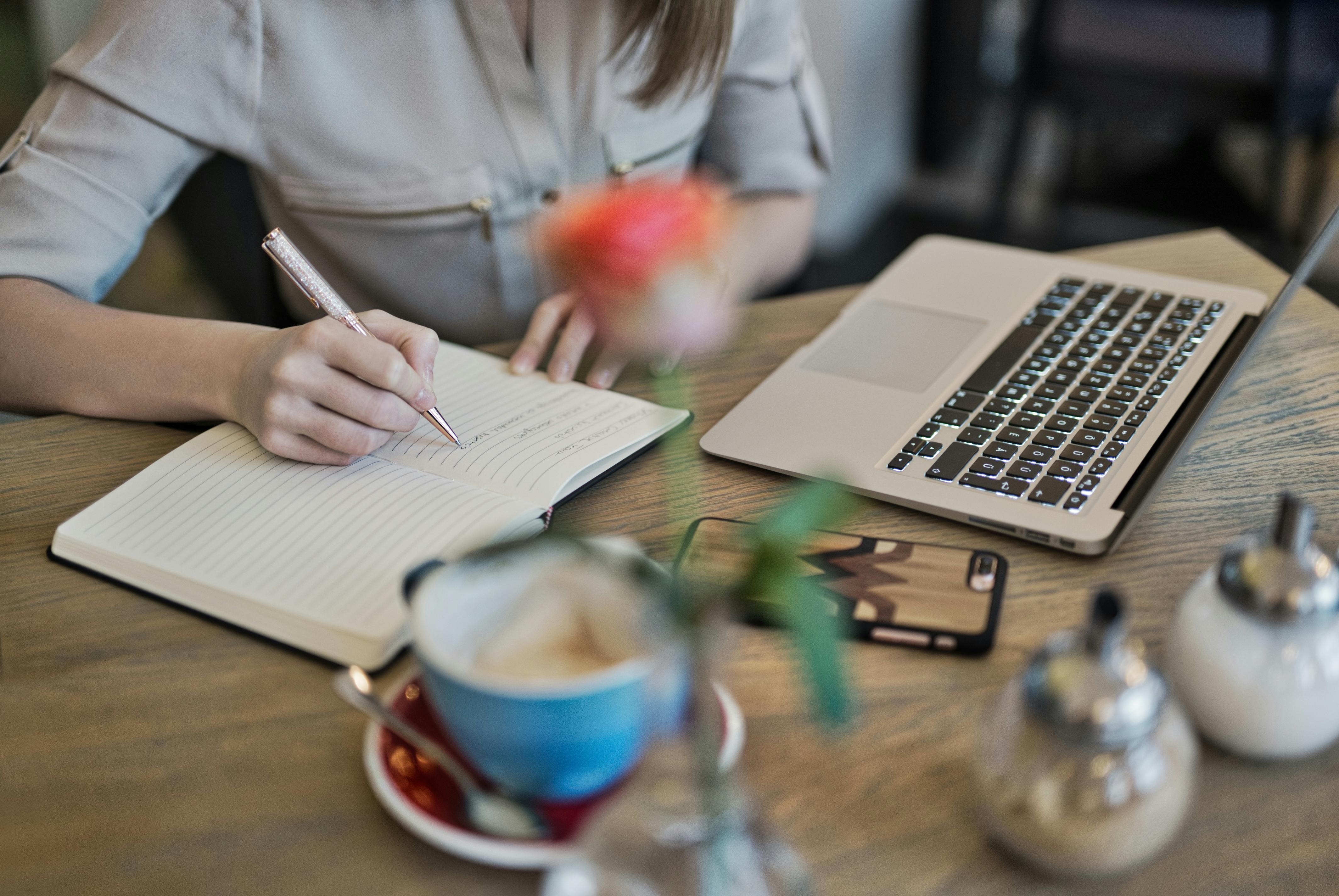 Person writing notes in a notebook beside a laptop and coffee, suggesting focused work or study in a café setting.