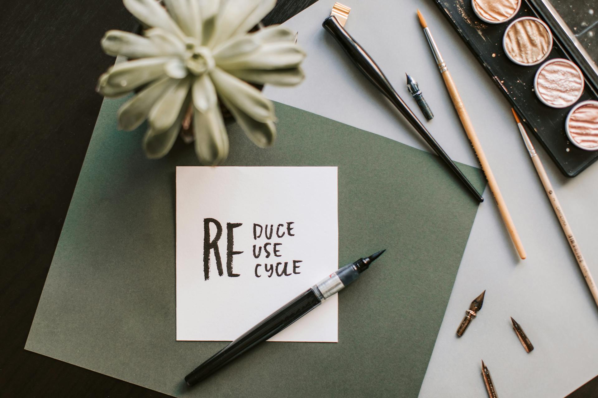 A handwritten note reading “Reduce, reuse, recycle” surrounded by art and writing tools on a desk.