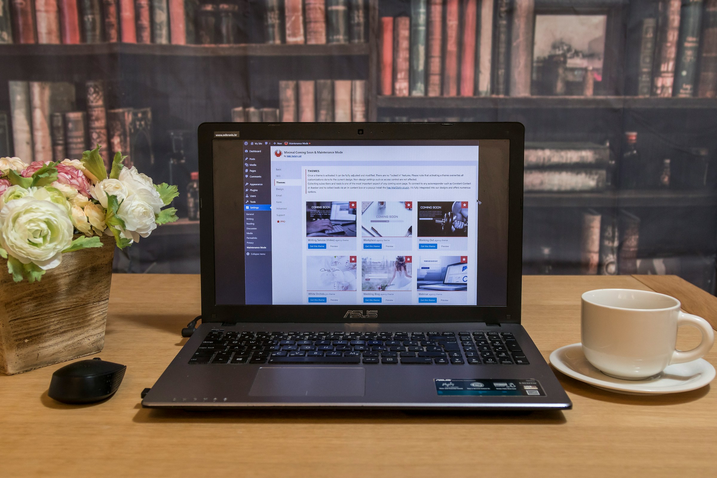 Laptop on a desk displaying a website dashboard, with a coffee cup and flowers.