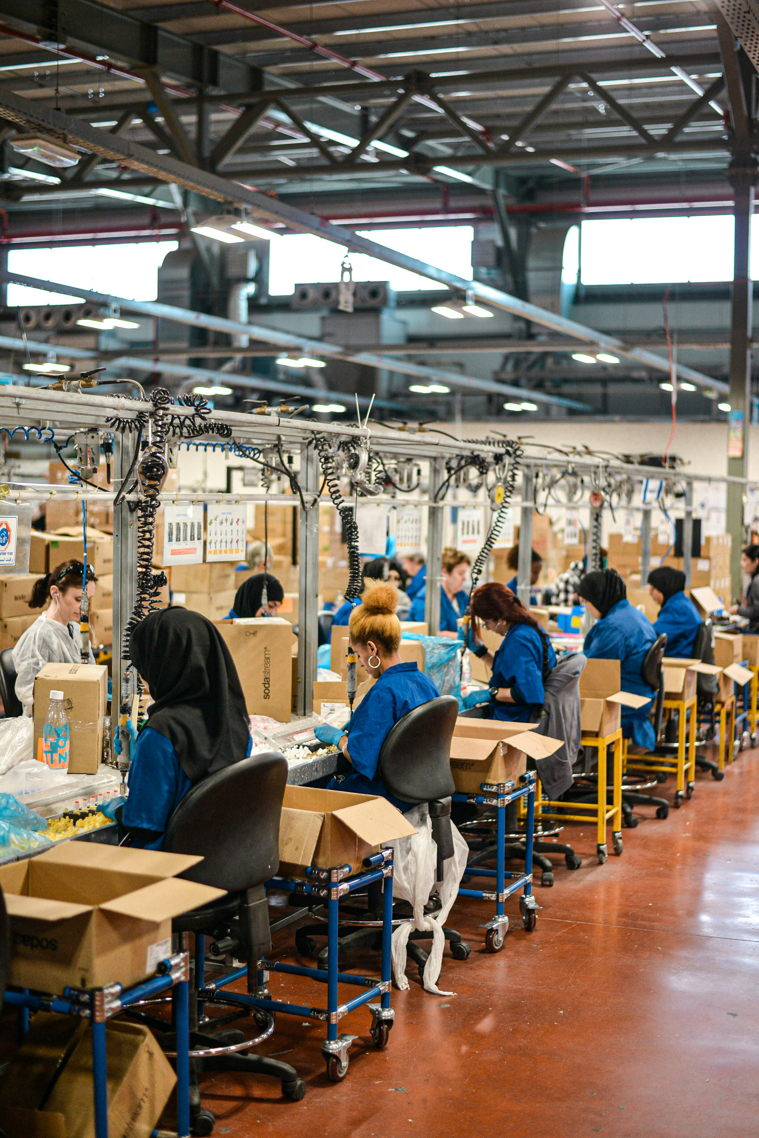 Female workers seated along an assembly line in a large factory next to cardboard boxes. They wear blue uniforms and sit at individual stations with tools and hanging cables overhead inside an industrial warehouse.