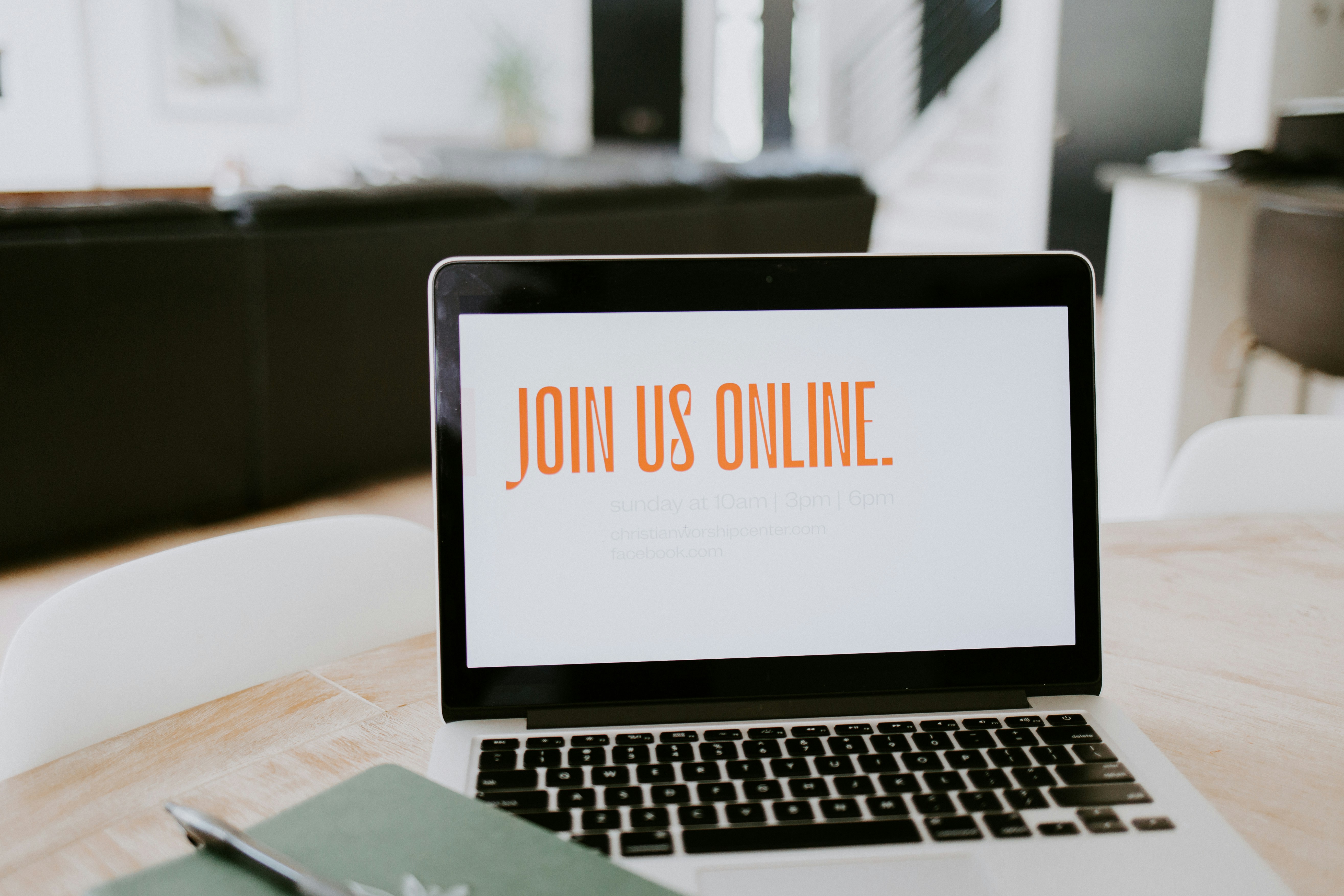 Colour photograph of an open laptop on a wooden table, with “Join us online” on the screen and a personal notepad next to the keyboard. Photograph by Samatha Borges on unsplash.org