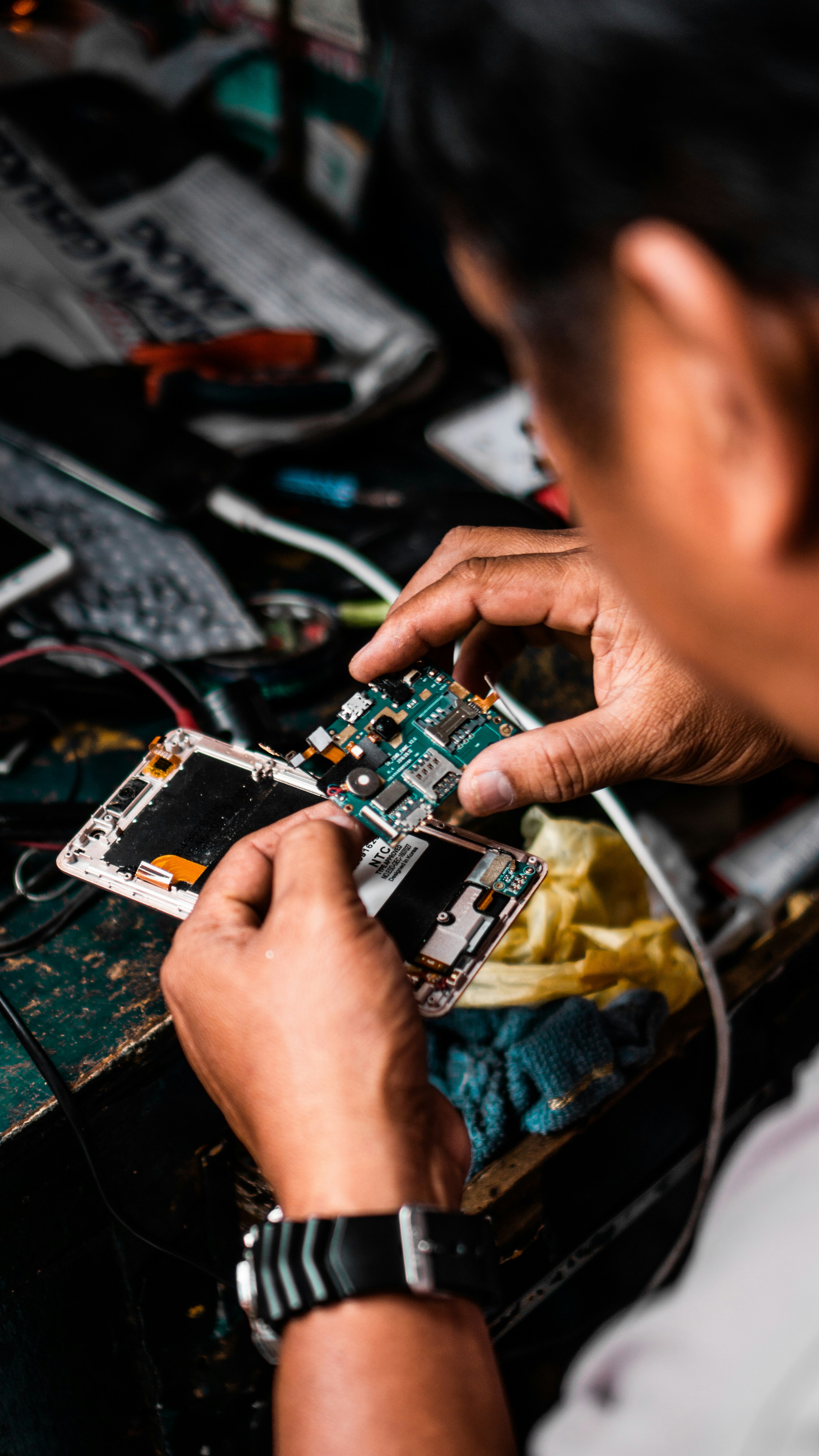Person repairing a device, holding its internal circuit board over an open device on a cluttered desk