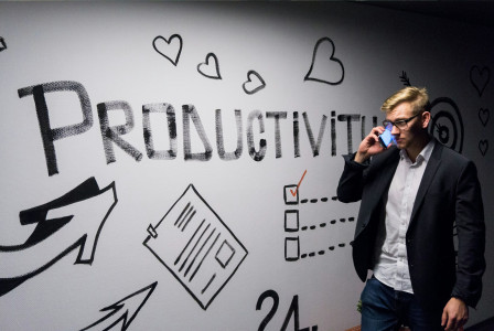 Colour photograph of man walking in front of a white wall with the word "productivity" on it while speaking on the phone, by Andreas Klassen on unsplash.