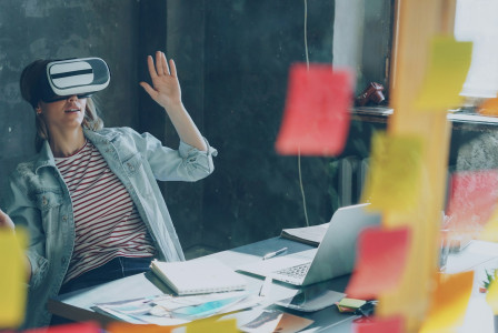 Colour photograph of woman wearing a vr headset while at her desk with laptop and sticky notes, by Vitaly Gariev on unsplash. 
