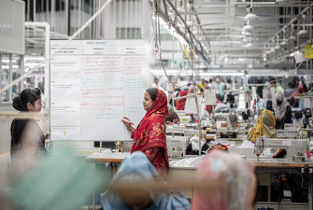 A woman in a red headscarf and a woman in a black dress review notes on a whiteboard in a busy garment production floor.