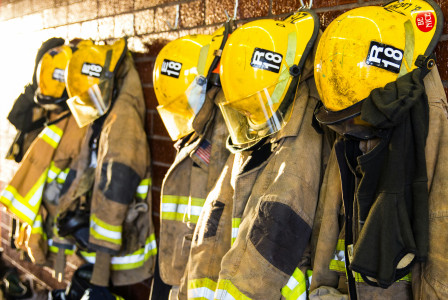 Several yellow firefighter helmets and protective turnout jackets hang on hooks along a brick wall in a fire station.
