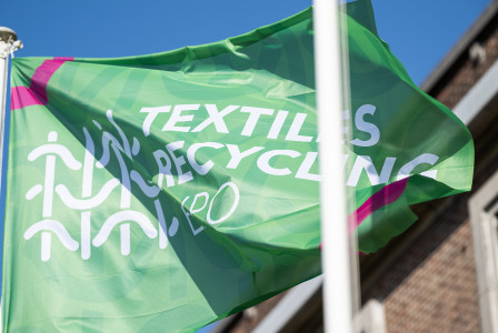Green “Textiles Recycling Expo” flag billowing in the wind against a bright blue sky, partially obscured by a white flagpole, with a brick building in the background.