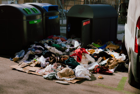 A photo presenting a pile of discarded clothing spilling out beside recycling bins on a street.