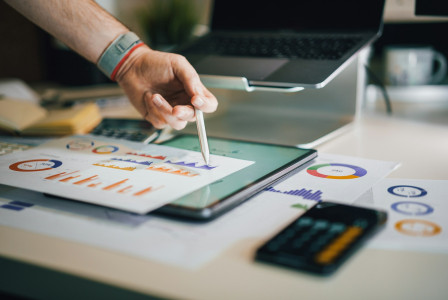 The photo presents a person analysing charts and financial data with a tablet, laptop and calculator.