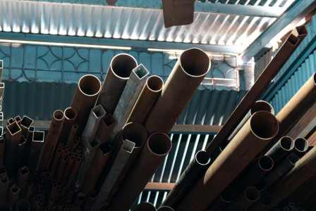 The picture describes steel pipes and metal profiles stored in an industrial warehouse, representing construction materials commonly used in building and infrastructure projects.