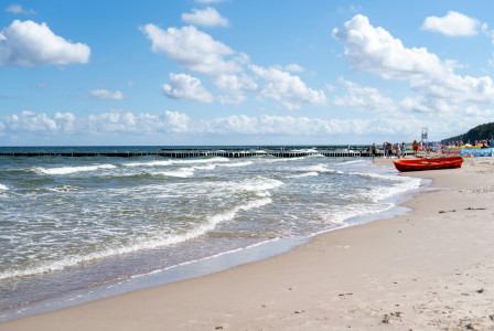 The photo presents waves washing onto a sandy beach, with a red boat and beachgoers under a blue sky with white clouds.