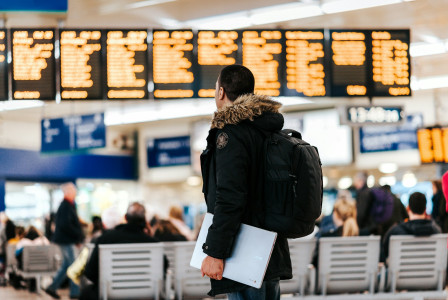 The photo presents a traveller with a backpack and laptop looking at departure boards in a busy terminal.