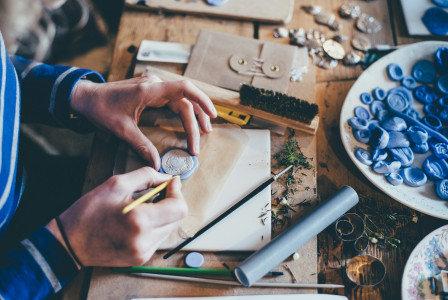 The photo presents a person crafting a small decorative piece at a wooden table covered with tools, moulds and plant cuttings.