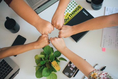 The photo presents five people bringing their fists together over a desk with laptops, notebooks, mugs and a potted plant.