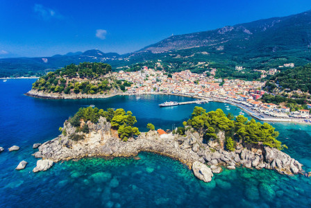 The photo presents an aerial view of a coastal town and harbour, with rocky, tree-covered islets in turquoise water and forested mountains behind.