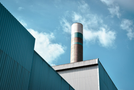The photo presents an industrial chimney rising above factory buildings under a blue sky.