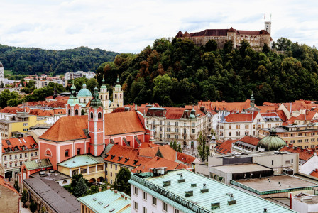 The photo presents a city with red-roofed buildings and church towers beneath a castle on a wooded hill.
