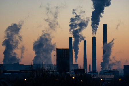 The photo presents an industrial facility with tall chimneys and large plumes rising into an orange sky.