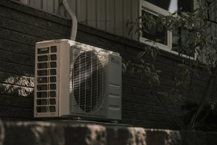 The photo presents an outdoor air-conditioning unit mounted beside a dark brick wall, with a leafy plant and window in the background.