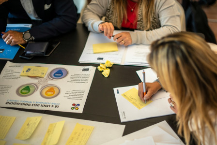 The photo presents people taking part in a workshop, writing on yellow sticky notes around a table covered with printed materials.