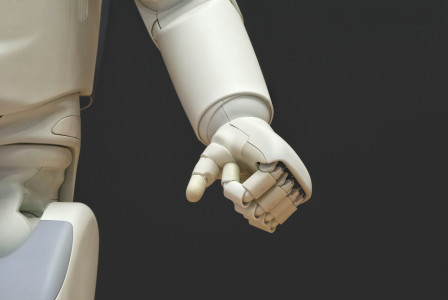The photo presents a close-up of a white humanoid robot’s arm and articulated hand against a dark background.