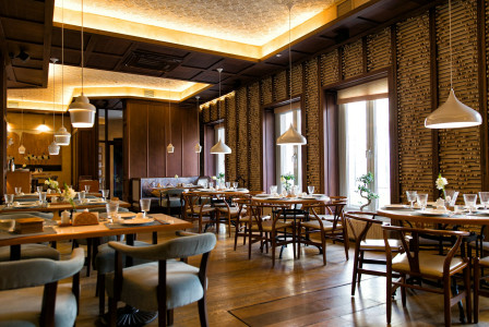 The photo presents an empty restaurant with set dining tables, wooden chairs, white pendant lights and large windows, surrounded by decorative wooden walls and warm lighting.