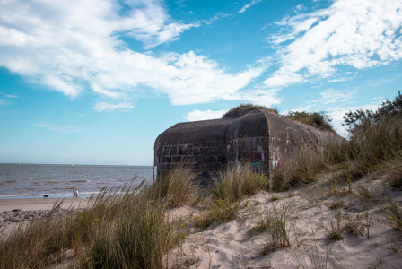 The photo presents a weathered concrete bunker among grassy sand dunes beside the sea, with two people and a dog walking along the beach.