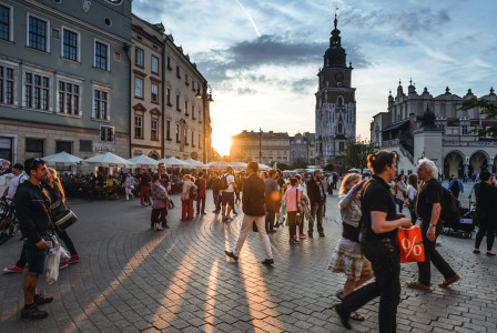 The photo presents a busy city square at sunset, with people walking past outdoor cafés, historic buildings and a tall clock tower.