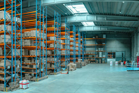 The photo presents a spacious warehouse with tall blue-and-orange storage racks, stacked boxes, pallets, loading bays and marked floor areas.