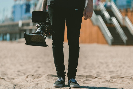 The photo presents a person carrying a professional video camera while standing on a sandy surface near an escalator.