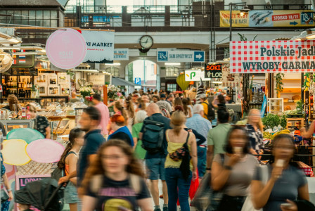 The photo presents a crowded indoor market with people walking between stalls selling food, drinks, flowers and fresh produce.