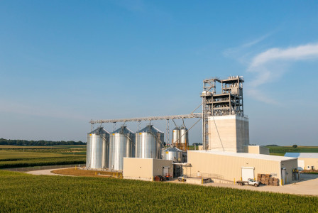 The photo presents a grain processing facility with tall metal silos and a large industrial tower surrounded by agricultural fields.