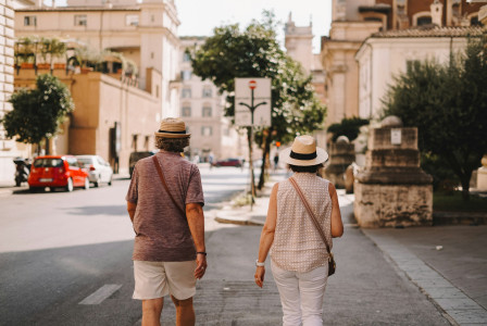 The photo presents two people wearing straw hats walking along a sunny street lined with historic buildings and parked cars.