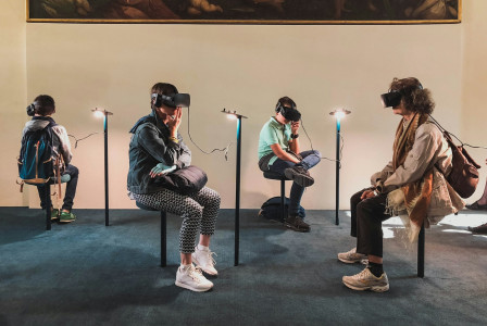 Four visitors wearing virtual reality headsets while seated in a museum or gallery space, with a large painting displayed on the wall above them.