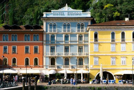 The photo presents colourful waterfront buildings, including Hotel Europa, with outdoor cafés and people along the promenade beneath a wooded hillside.
