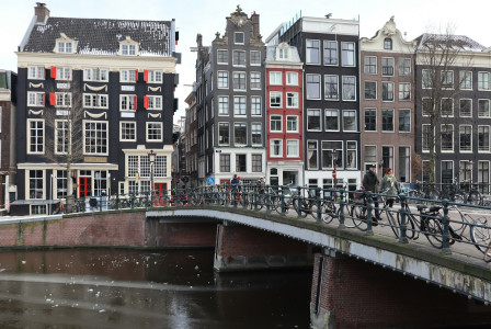 The photo presents people crossing a canal bridge lined with bicycles, with traditional canal houses in the background.