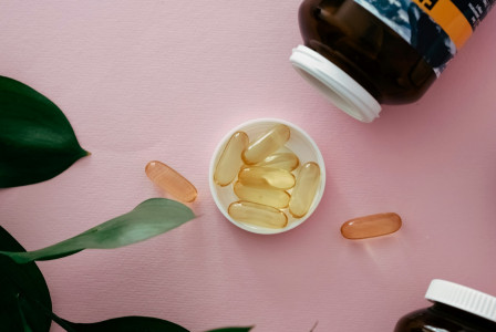 The photo presents golden supplement capsules in a white bottle cap, with loose capsules, brown bottles and green leaves on a pink background.