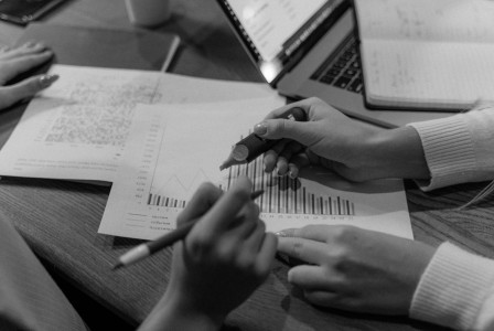 Monochrome image of hands analysing documents and charts in an office setting.