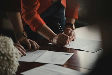 The photo presents a person signing a document at a wooden table, with other people and papers nearby.