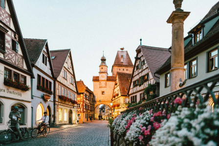 The photo presents a cobbled street lined with traditional timber-framed houses, flowers and a clock tower.