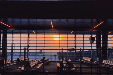 The photo presents an empty airport departure lounge with rows of seats and large windows overlooking an aircraft and an orange sunset.
