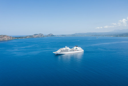 The photo presents a white cruise ship on calm blue water, with a rocky coastline and distant mountains beneath a clear sky.