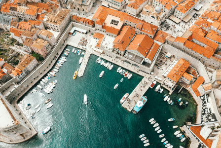 The photo presents an aerial view of a harbour filled with boats, surrounded by stone buildings with red-tiled roofs.